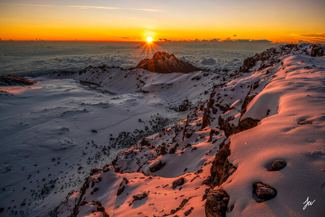 Mount Kilimanjaro summit sunrise with Mawenzi Peak illuminated beneath a dramatic sunburst, viewed from the upper slopes of Africa’s highest mountain.
