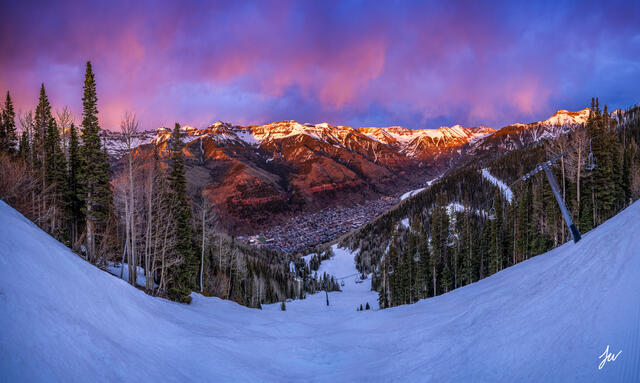 A panoramic winter sunset view of Telluride, Colorado showing the ski resort, mountain peaks, and town in the valley below.